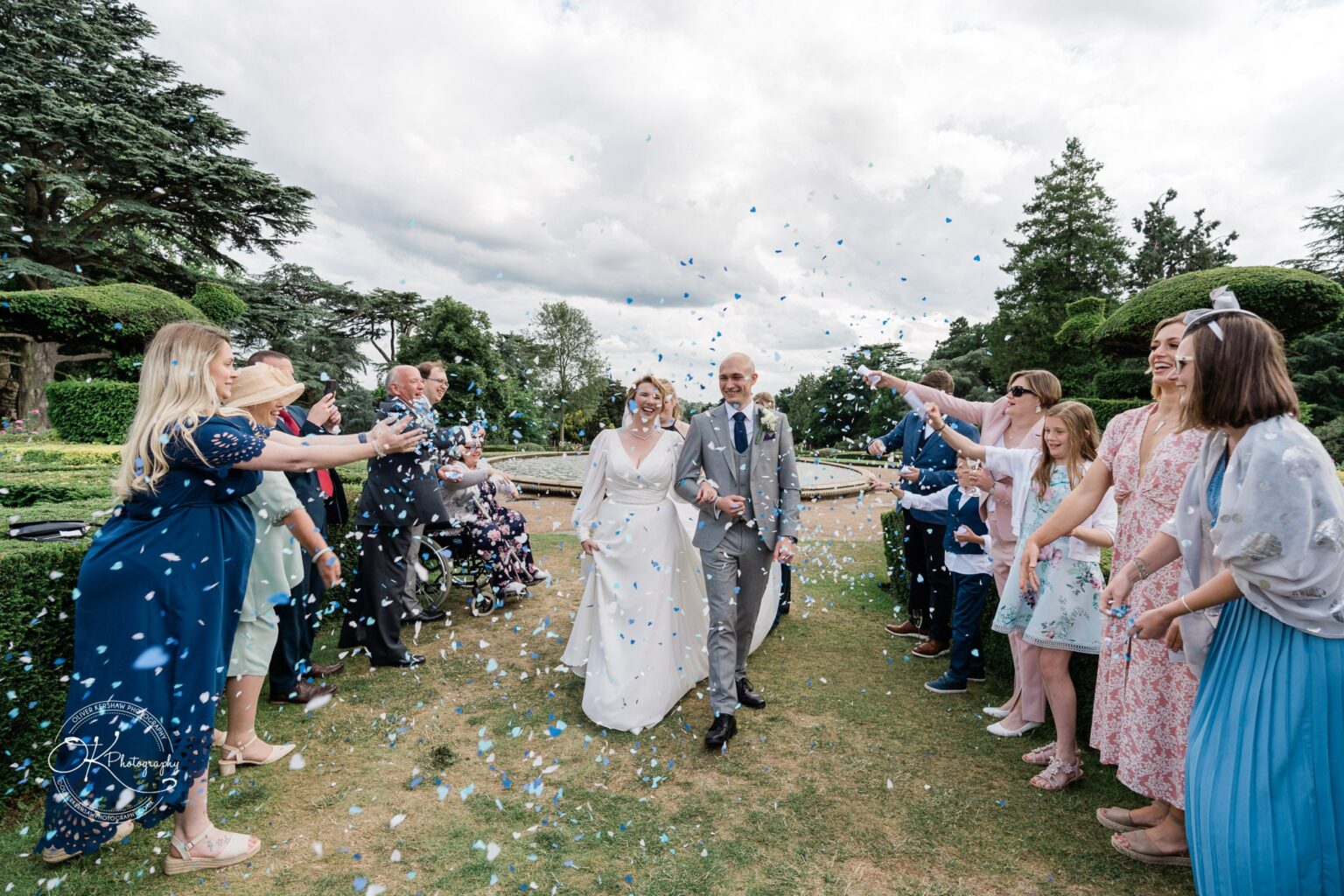 A newlywed couple walks hand in hand down an aisle while wedding guests toss blue confetti at them in a garden setting.