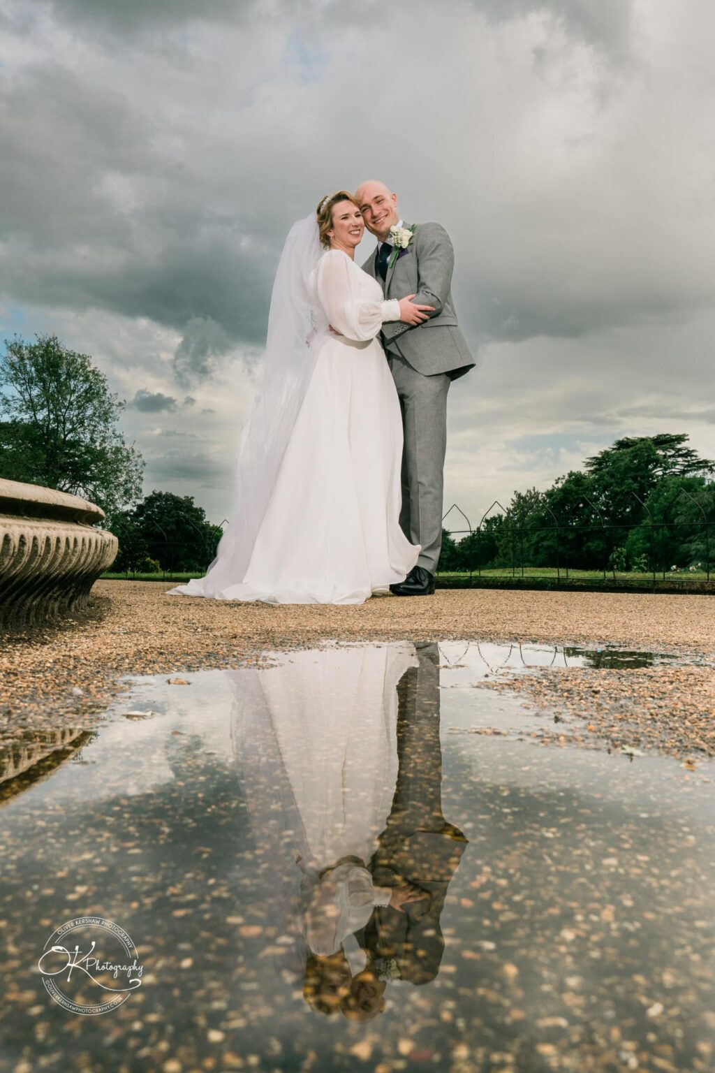 Bride and groom standing on a gravel path with reflections in a puddle, surrounded by greenery on a cloudy day.