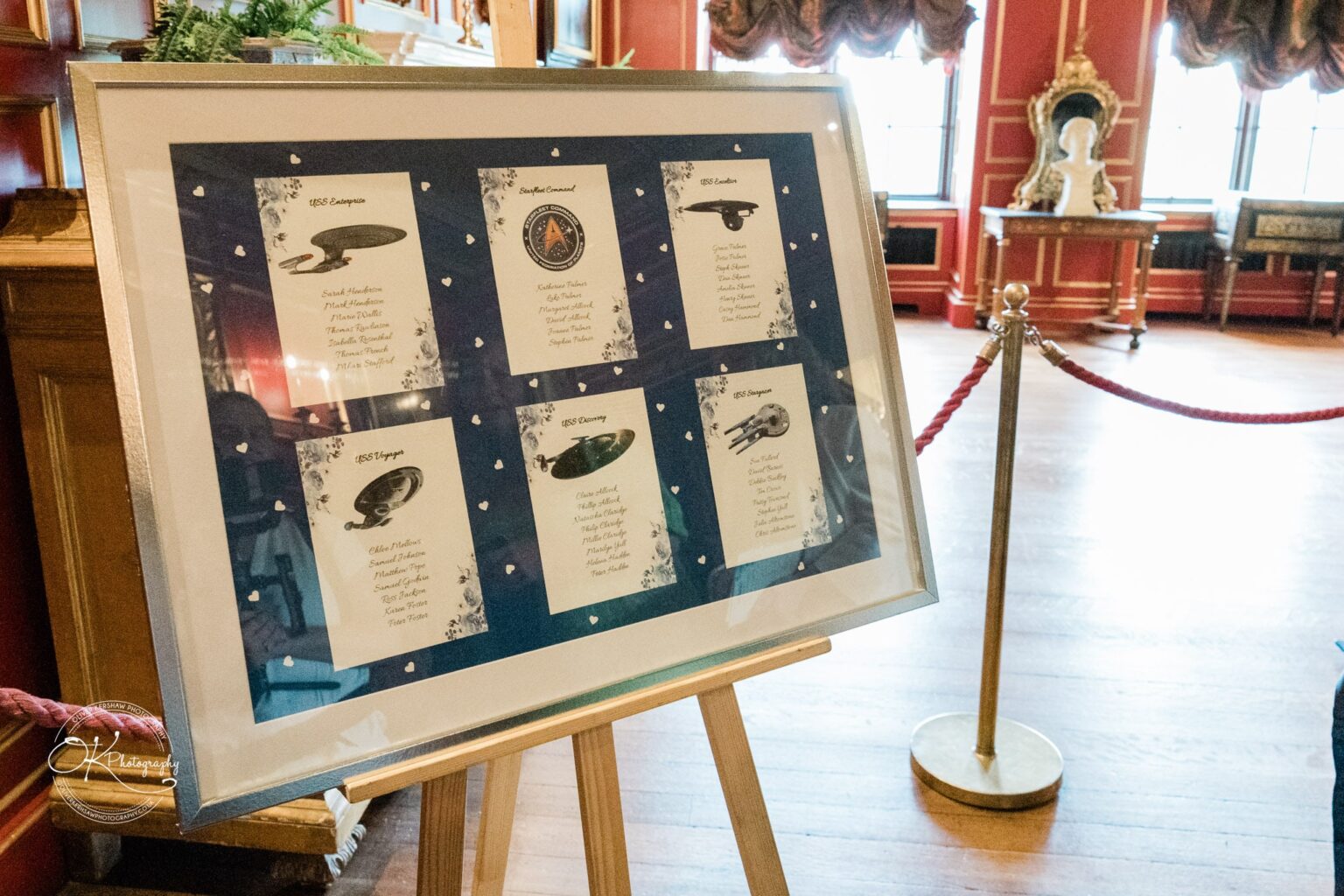 A framed display on an easel featuring Star Trek ship illustrations and crew lists in a room with ornate decor at Warwick Castle.