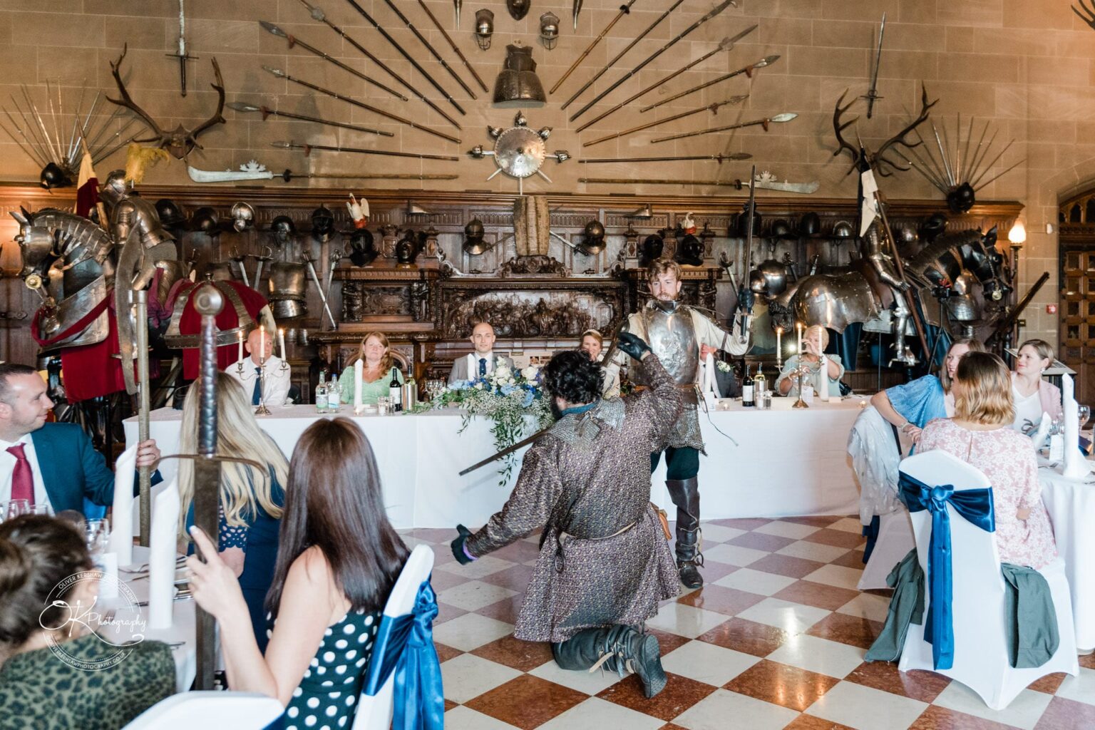 A medieval-themed event in a hall adorned with armour, weapons, and shields, featuring a long table with seated guests and two people in period costumes performing on the floor.