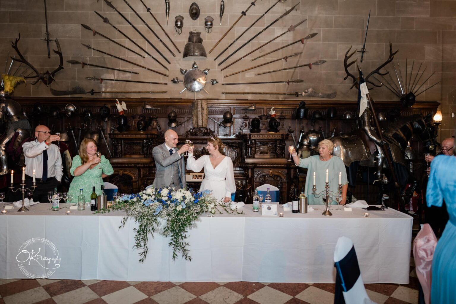 A wedding party toasting at a banquet table decorated with blue and white flowers, inside Warwick Castle with medieval armour and weapons displayed on the wall in the background.