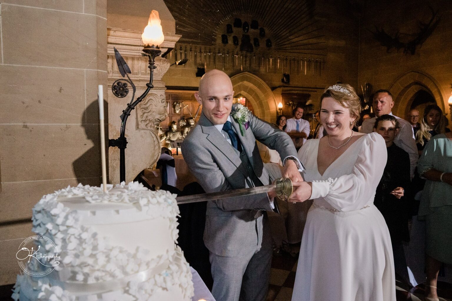 A bride and groom cut a white wedding cake with a sword in a medieval-themed room, surrounded by guests.