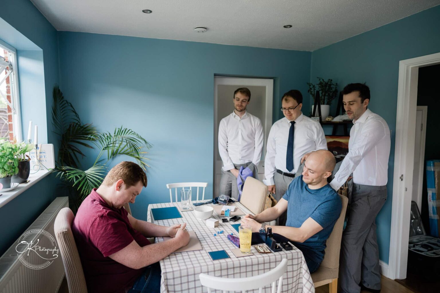 A group of men gather in a blue-painted dining room; two are seated at a table playing a game while three others stand watching.
