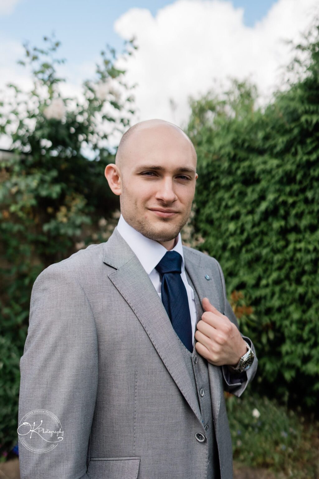 Man in a grey suit with a navy tie standing outside, with green foliage and a partly cloudy sky in the background.