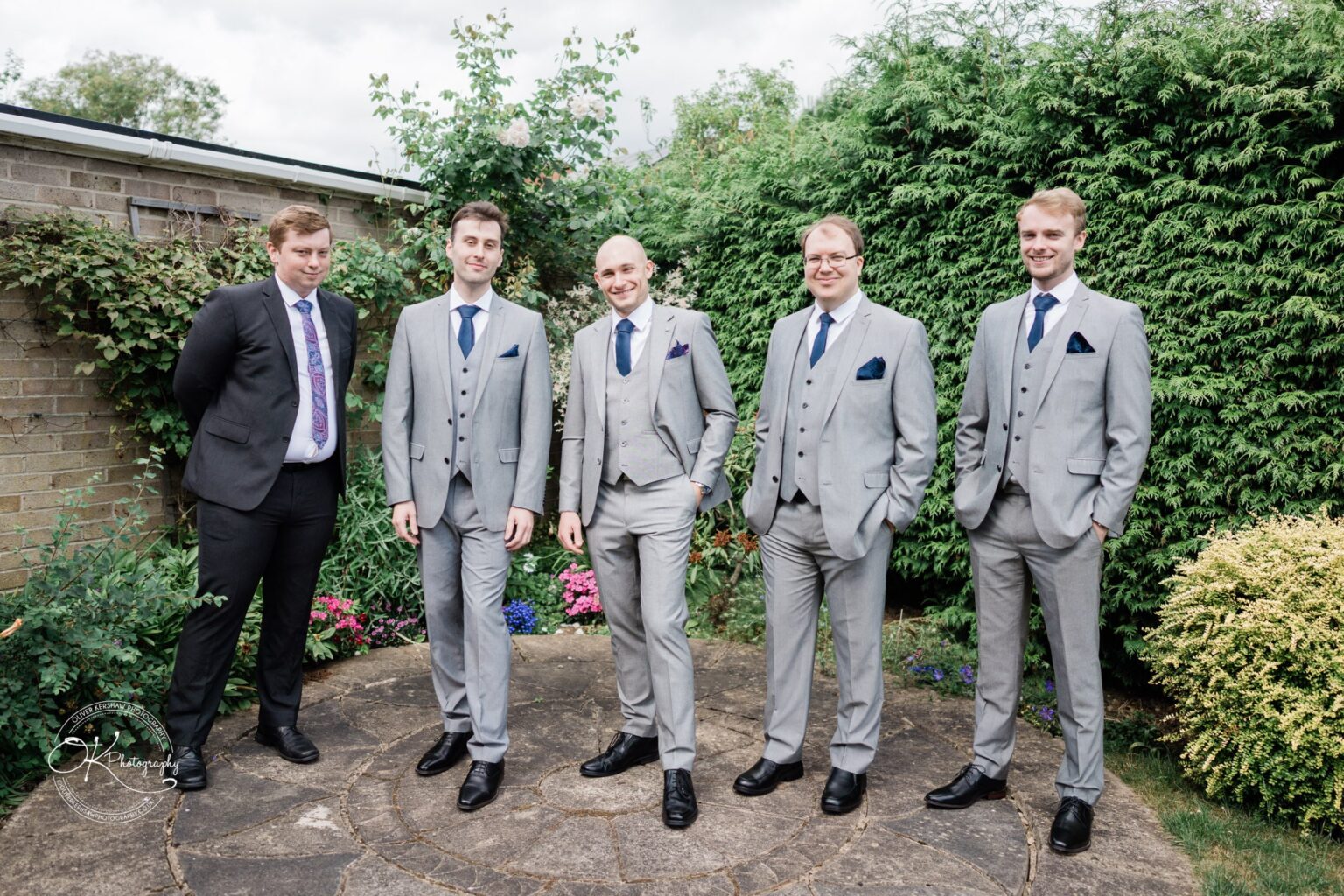 Five men in suits standing outdoors in front of greenery and flowers; four are in grey suits with blue ties and one in a black suit with a patterned tie.