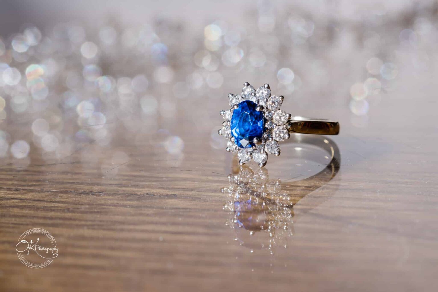 Gold ring with a blue sapphire surrounded by diamonds on a wooden surface with a blurred sparkly background.