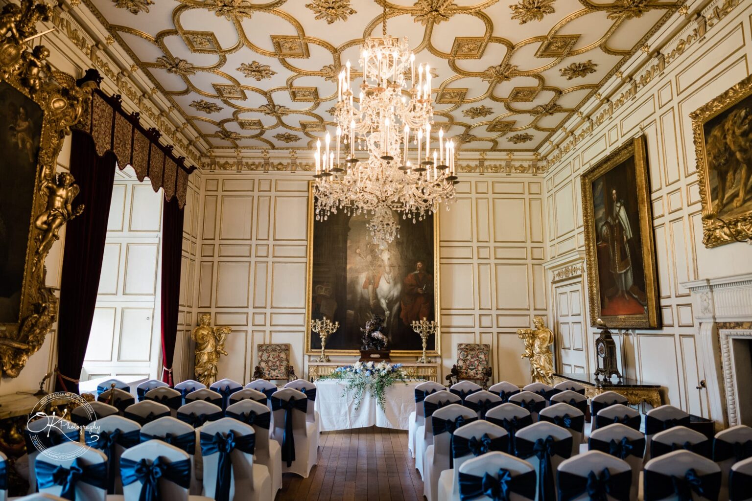 A grand hall at Warwick Castle set up for an event, featuring ornate golden ceiling details, chandeliers, paintings, and rows of chairs with white covers and navy blue bows.