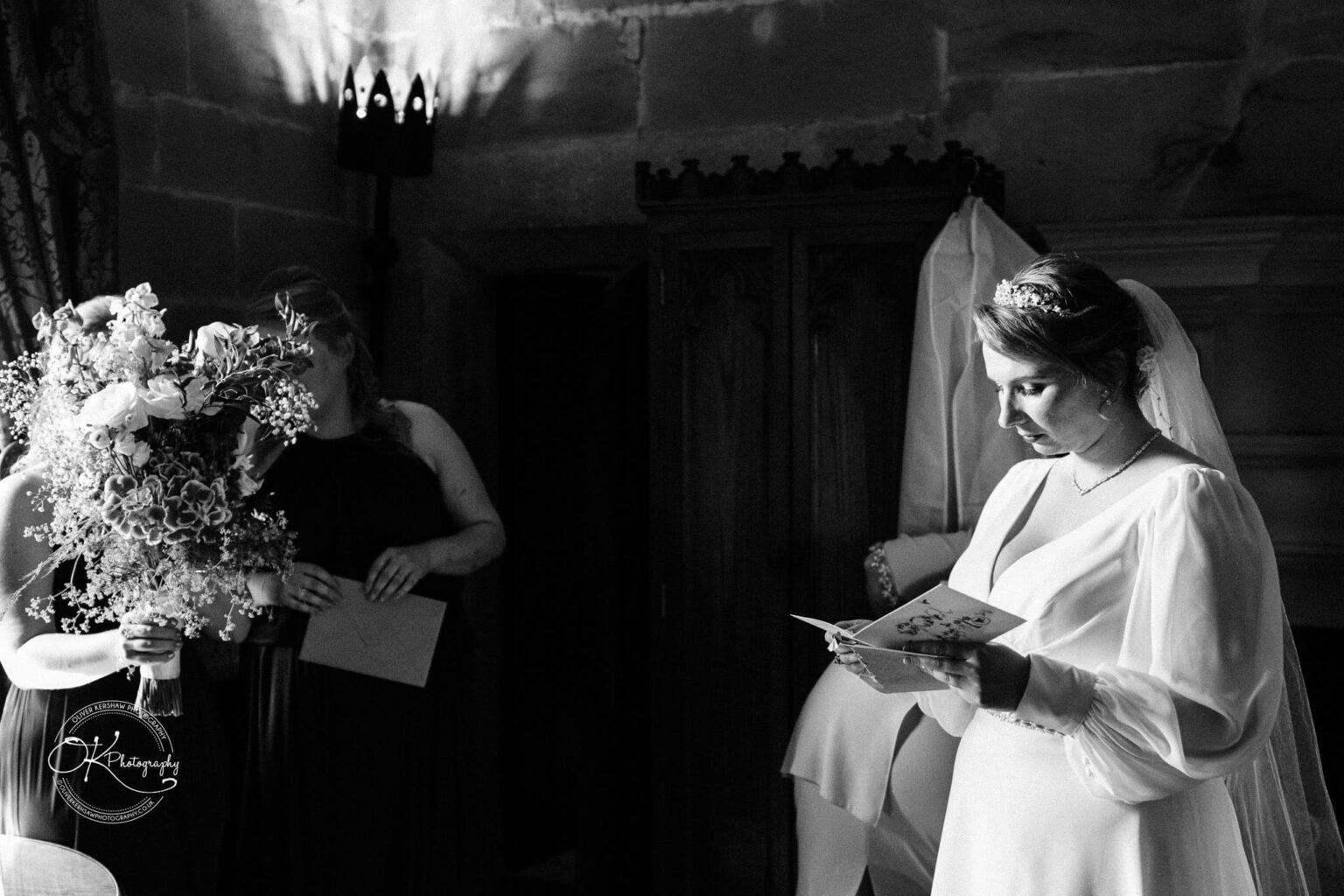 A bride in a white dress reads a card while another woman holds a large bouquet of flowers in a dimly lit room with stone walls.