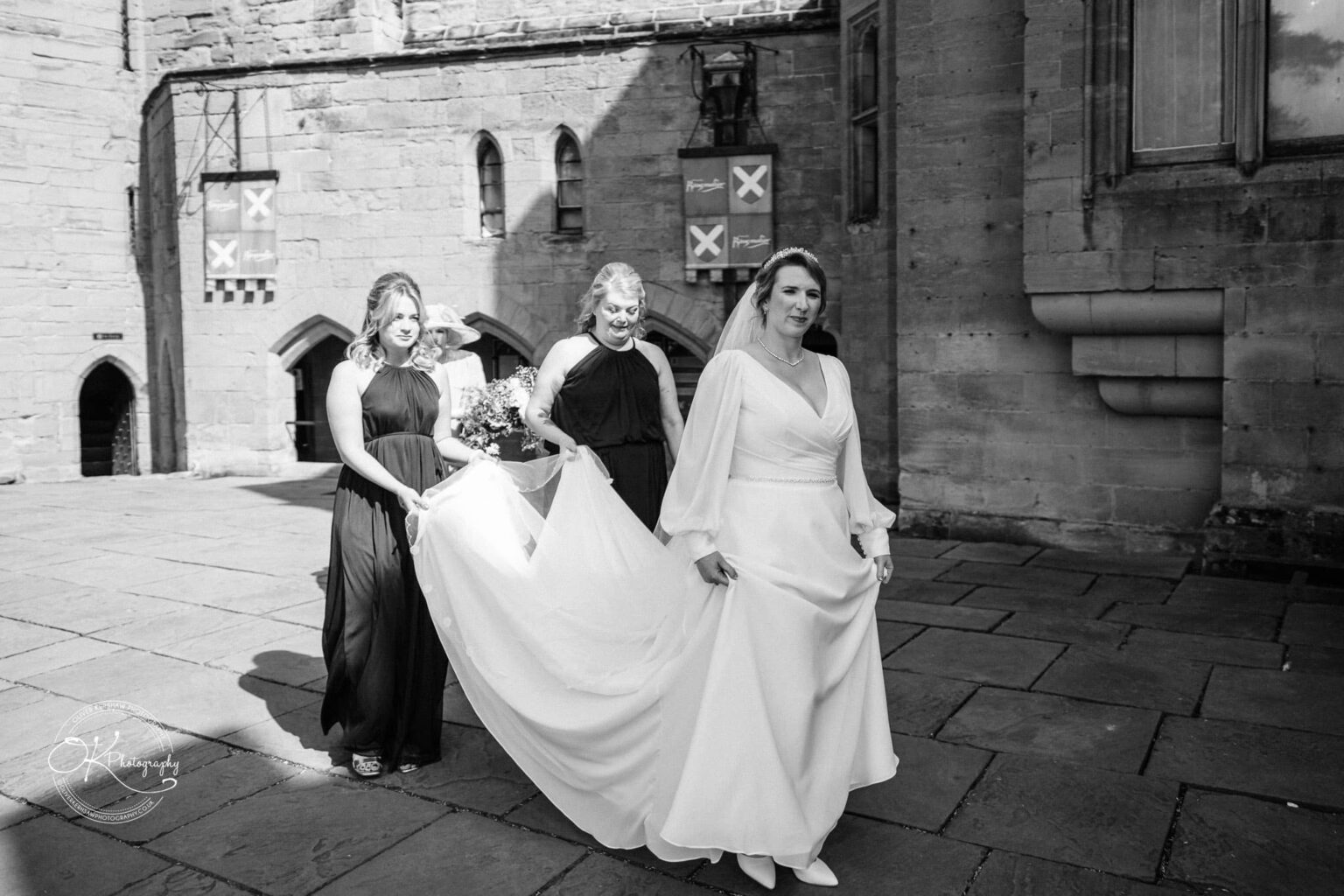 A black and white photo of a bride walking with her bridesmaids in front of a stone building, with a woman in a hat visible in the background.