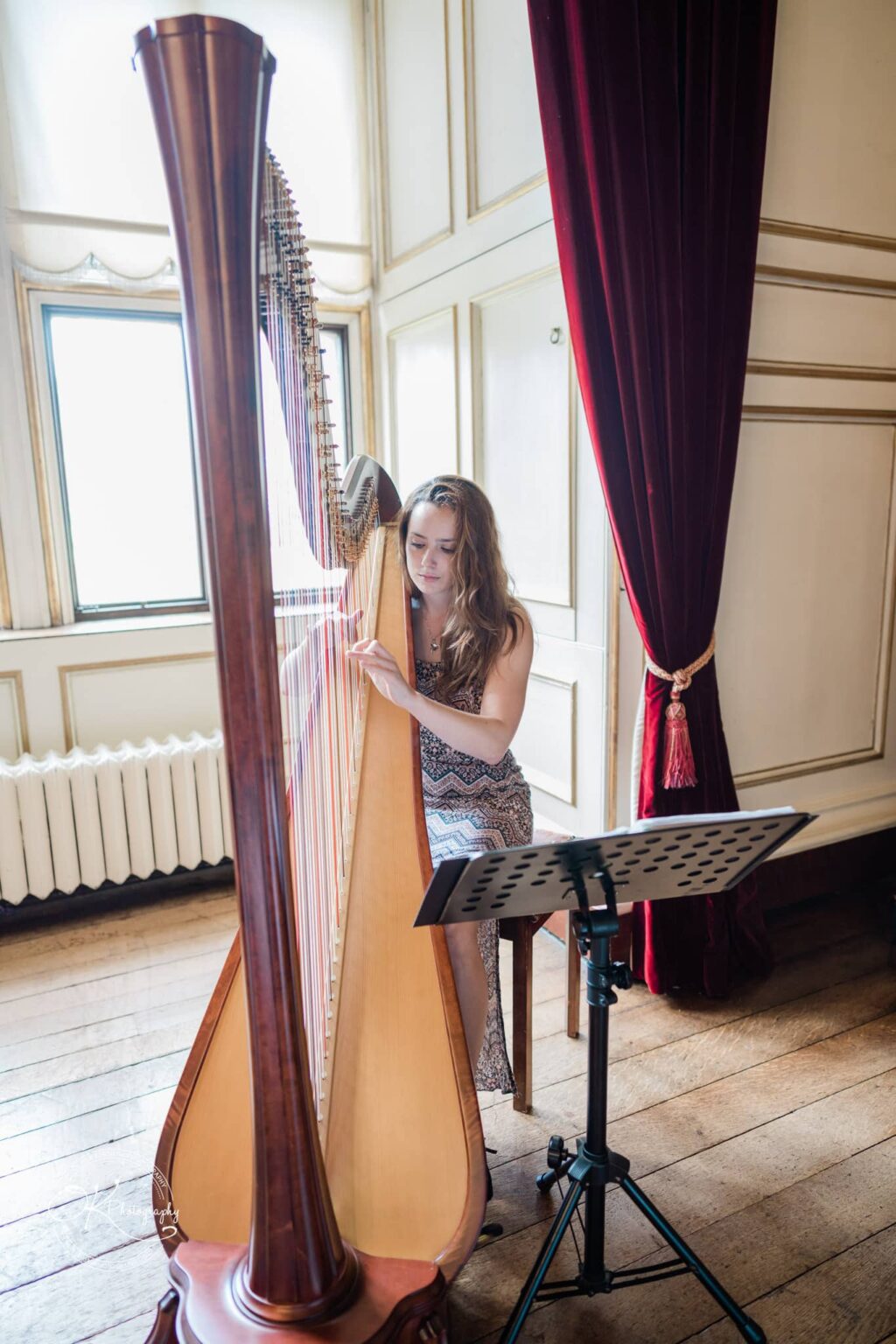 A woman playing a harp in an elegant room with wooden floors, red curtains, and large windows at Warwick Castle.