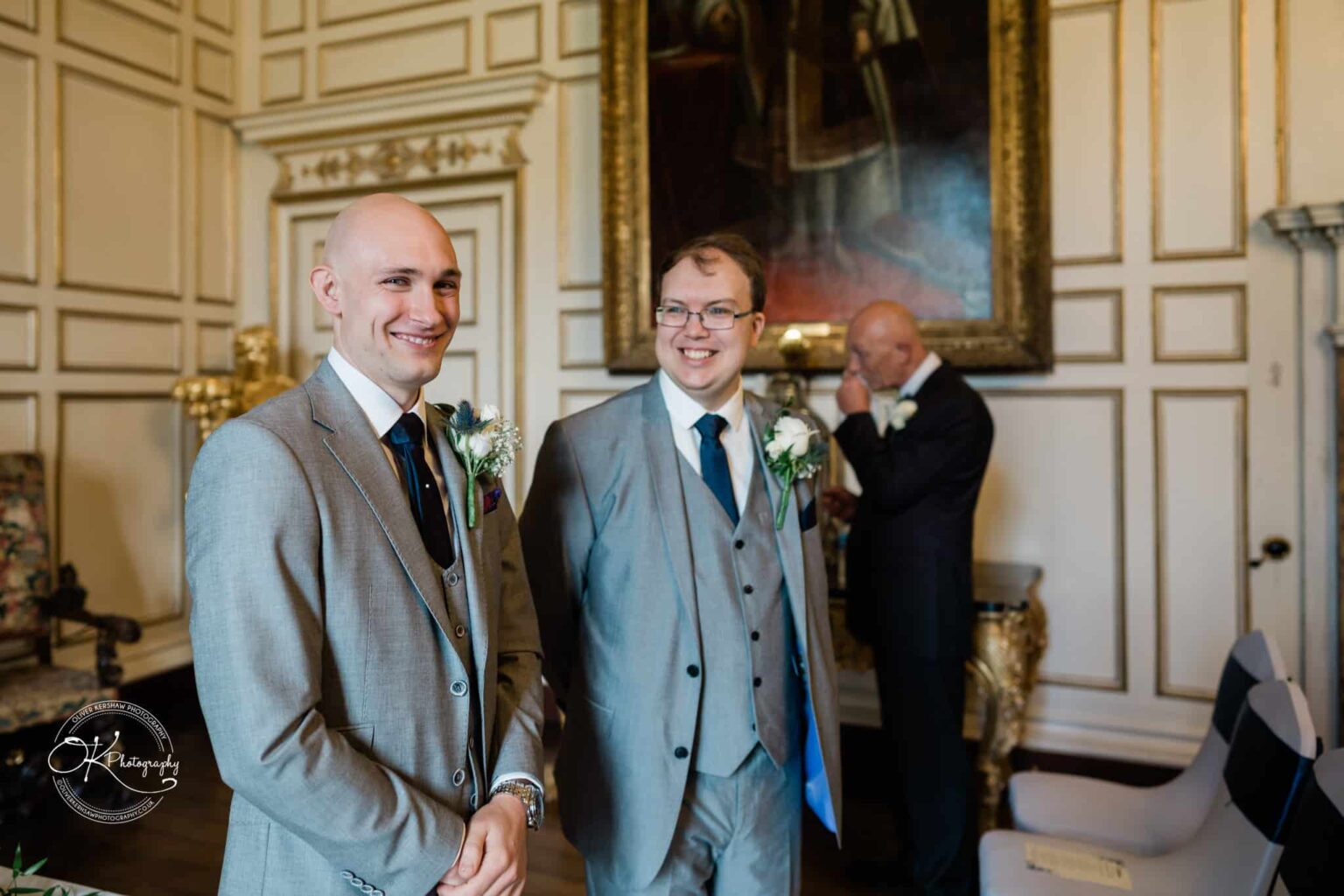 Two men in grey suits with boutonnieres smiling in an ornately decorated room, with another older man in the background adjusting his glasses.
