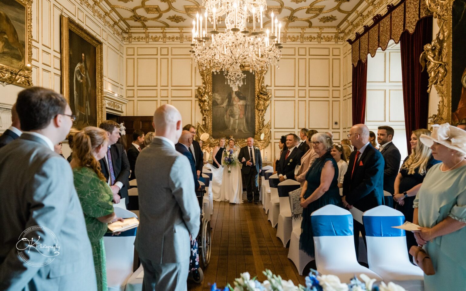 Bride walking down the aisle towards the groom in an ornate room filled with seated guests, chandeliers, and large paintings.