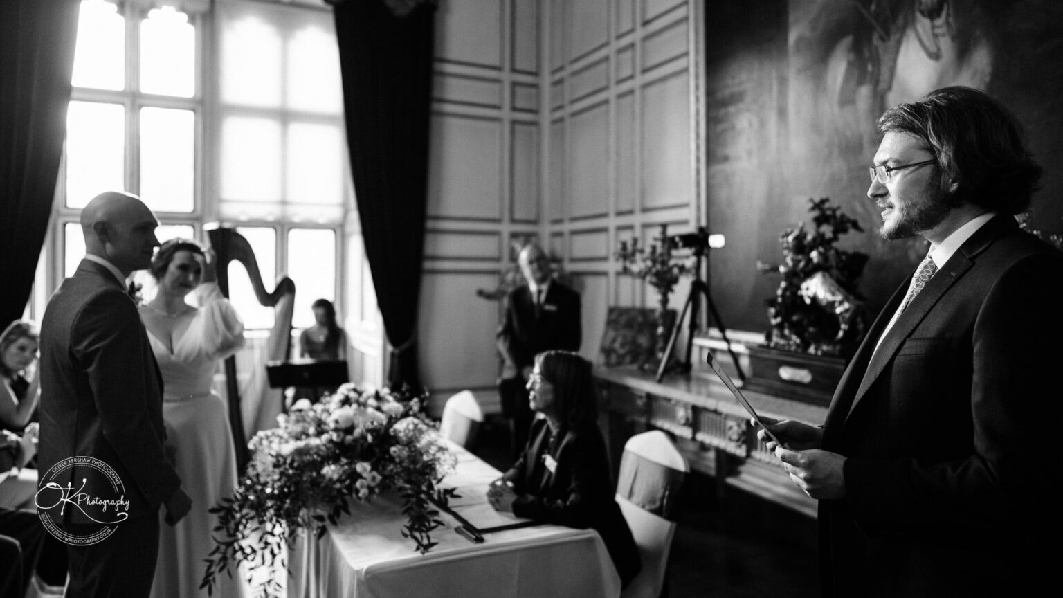 A wedding ceremony taking place indoors at Warwick Castle, featuring the couple standing in front of a table with floral arrangements, a harpist in the background, and a person delivering a speech.
