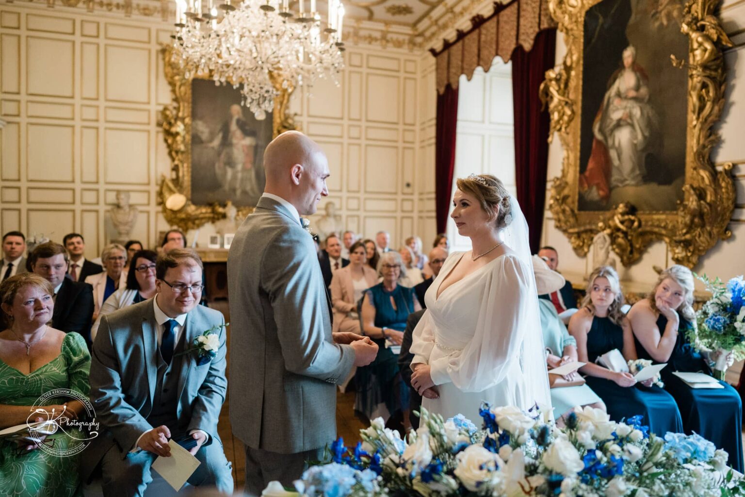 A couple exchanging vows in an ornate room decorated with chandeliers, large paintings, and white and blue flower arrangements, with guests seated and observing.