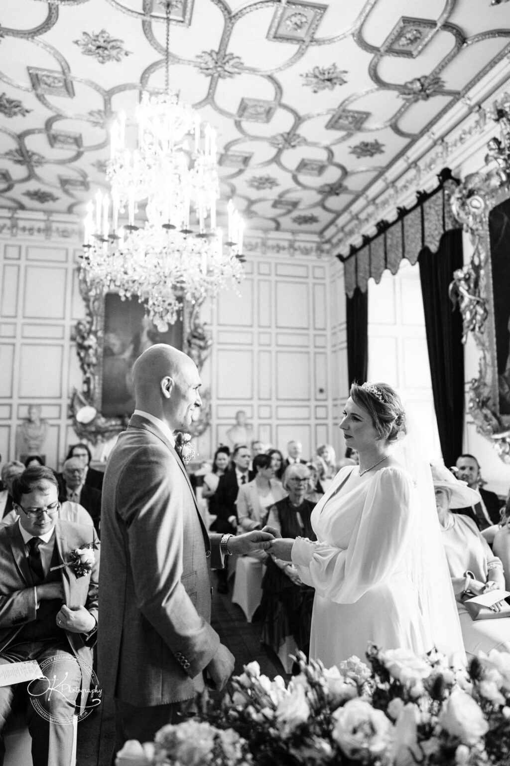 Black and white photo of a wedding ceremony in a grand, ornately decorated room with a large chandelier, paneled walls, and a crowd of seated guests, at Warwick Castle.