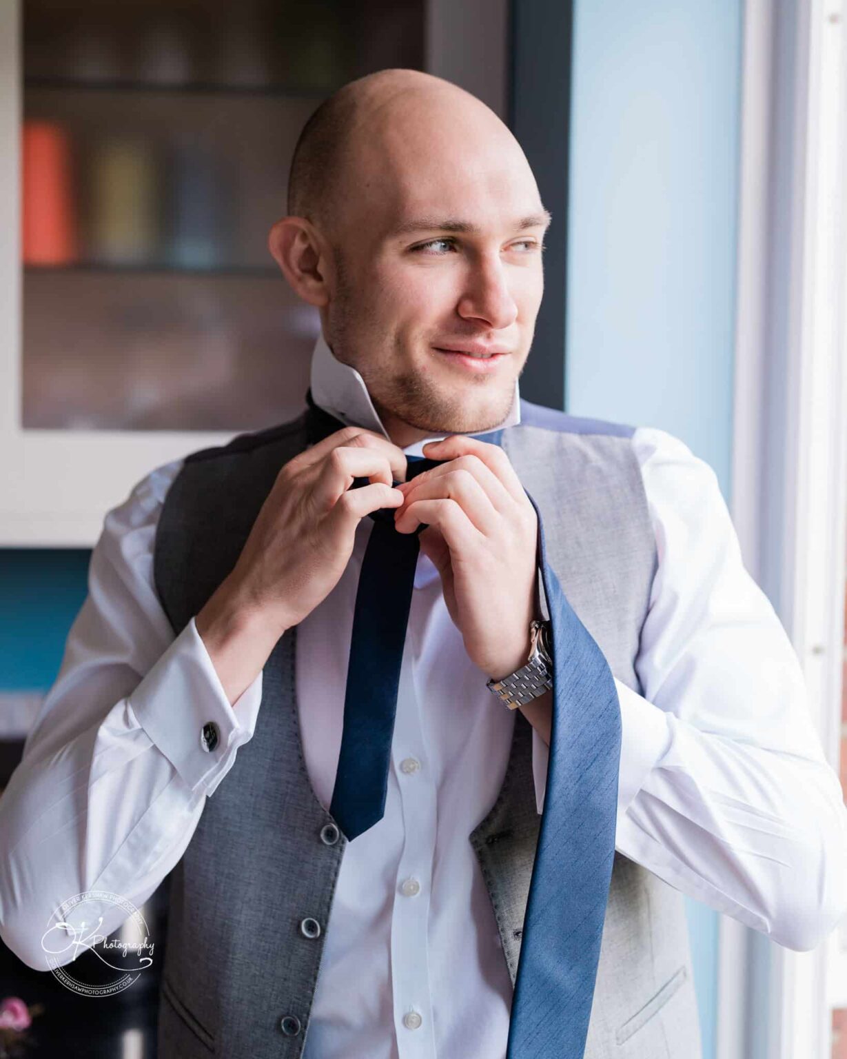 A man adjusting a dark blue tie while looking to the side, wearing a white dress shirt and grey vest.