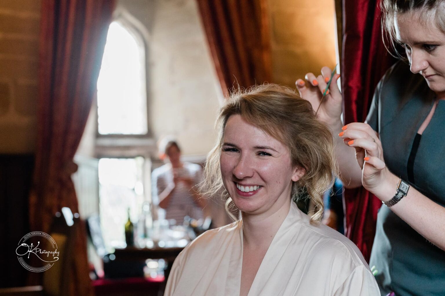 A woman smiling while her hair is being styled by another person in a room with a large window and red curtains.