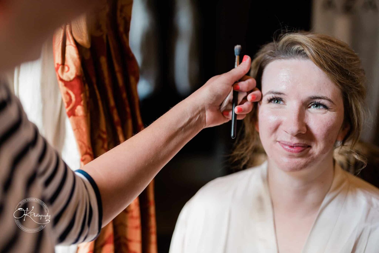 A smiling woman having her makeup done by another person holding a brush.
