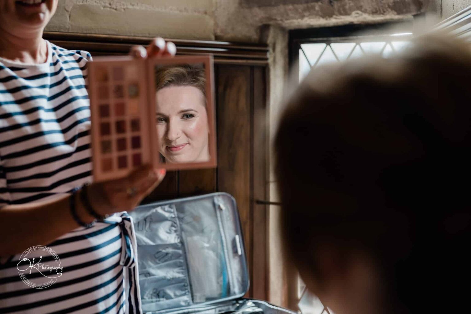 A woman holding an eyeshadow palette with a built-in mirror, reflecting the smiling face of another woman.