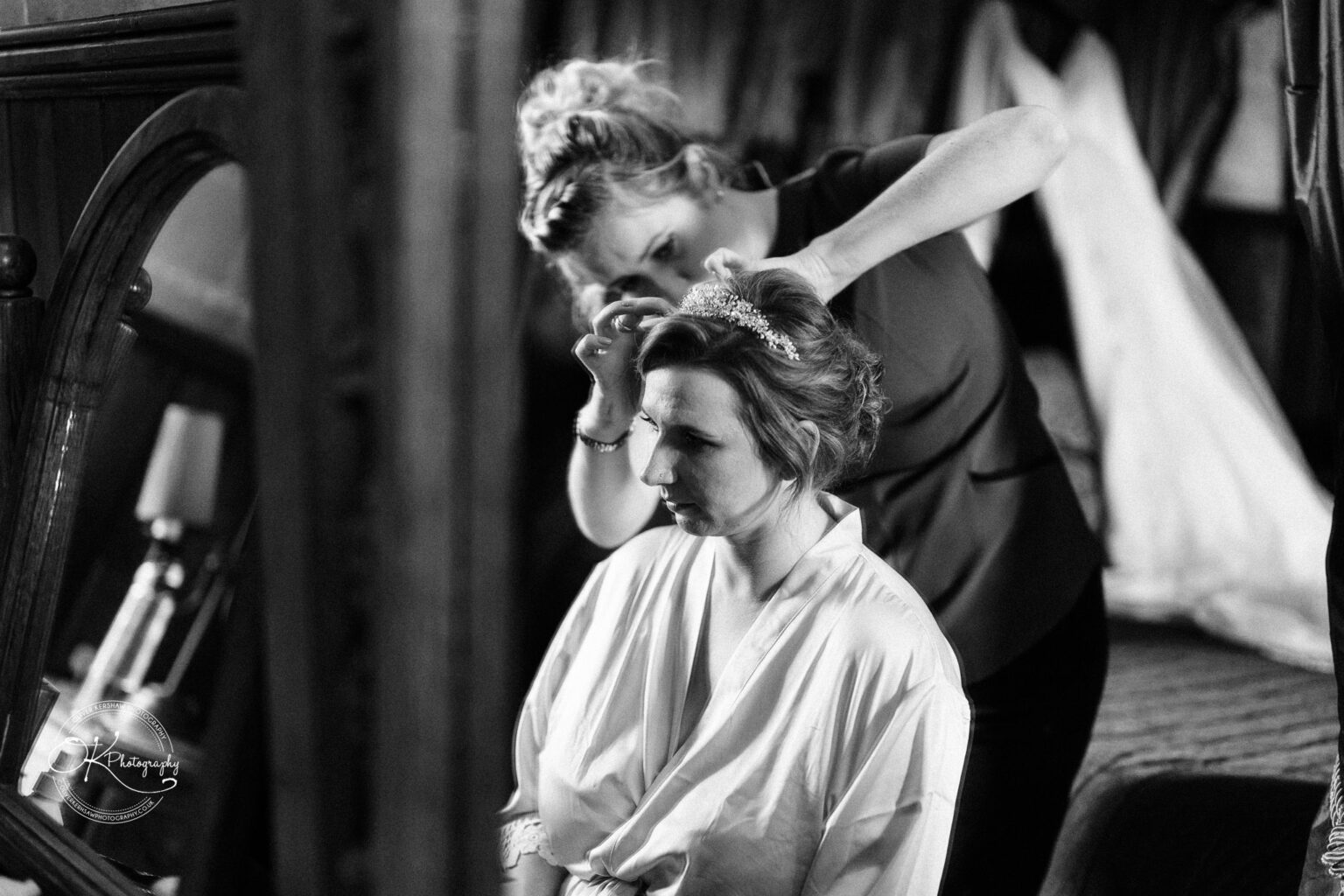 A woman gets her hair styled and crown adjusted by another woman in a dimly lit room.