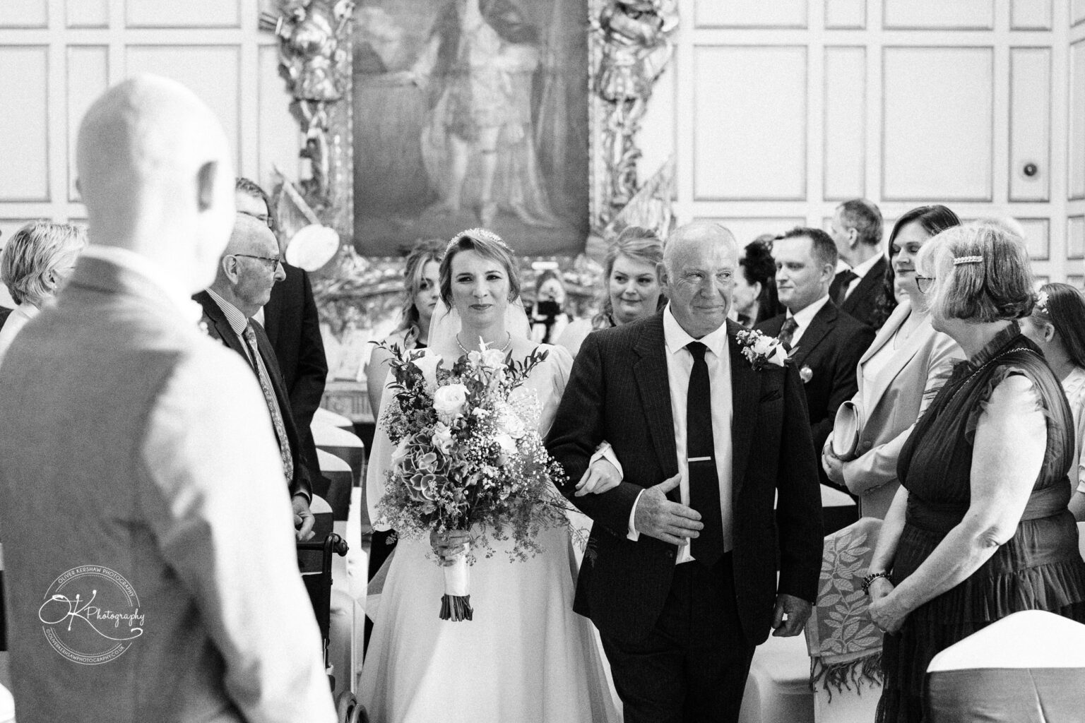 A bride and her father walk down the aisle at a wedding ceremony, surrounded by guests, in a historical room at Warwick Castle.