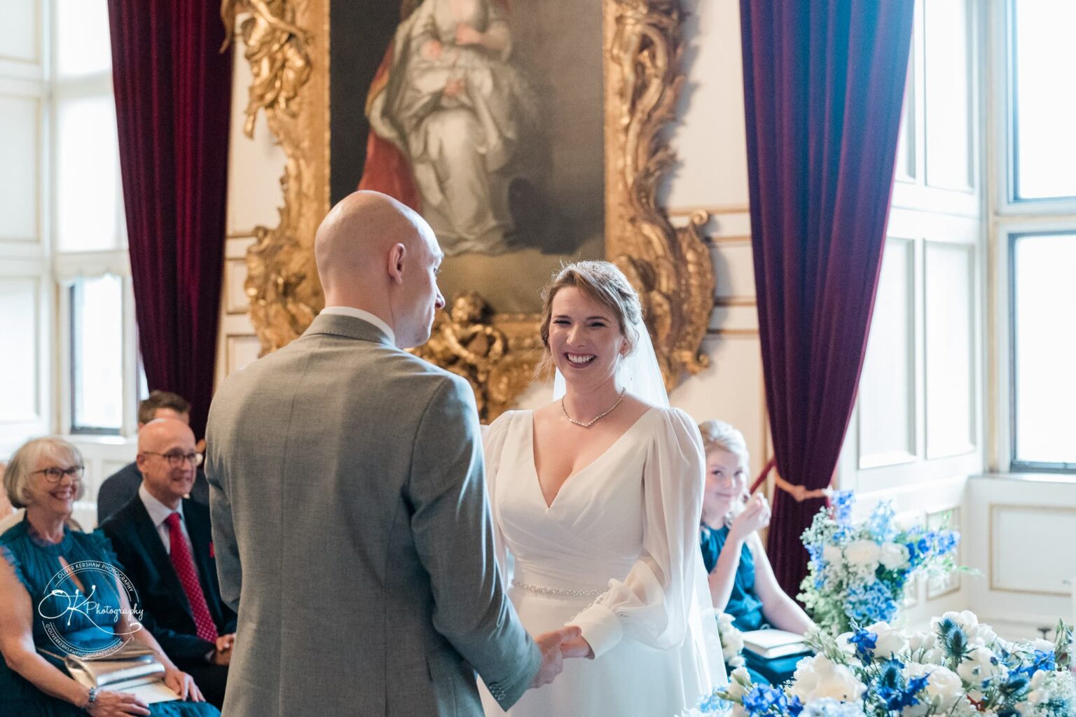 A bride and groom standing and holding hands during their wedding ceremony, with guests seated and observing in the background.