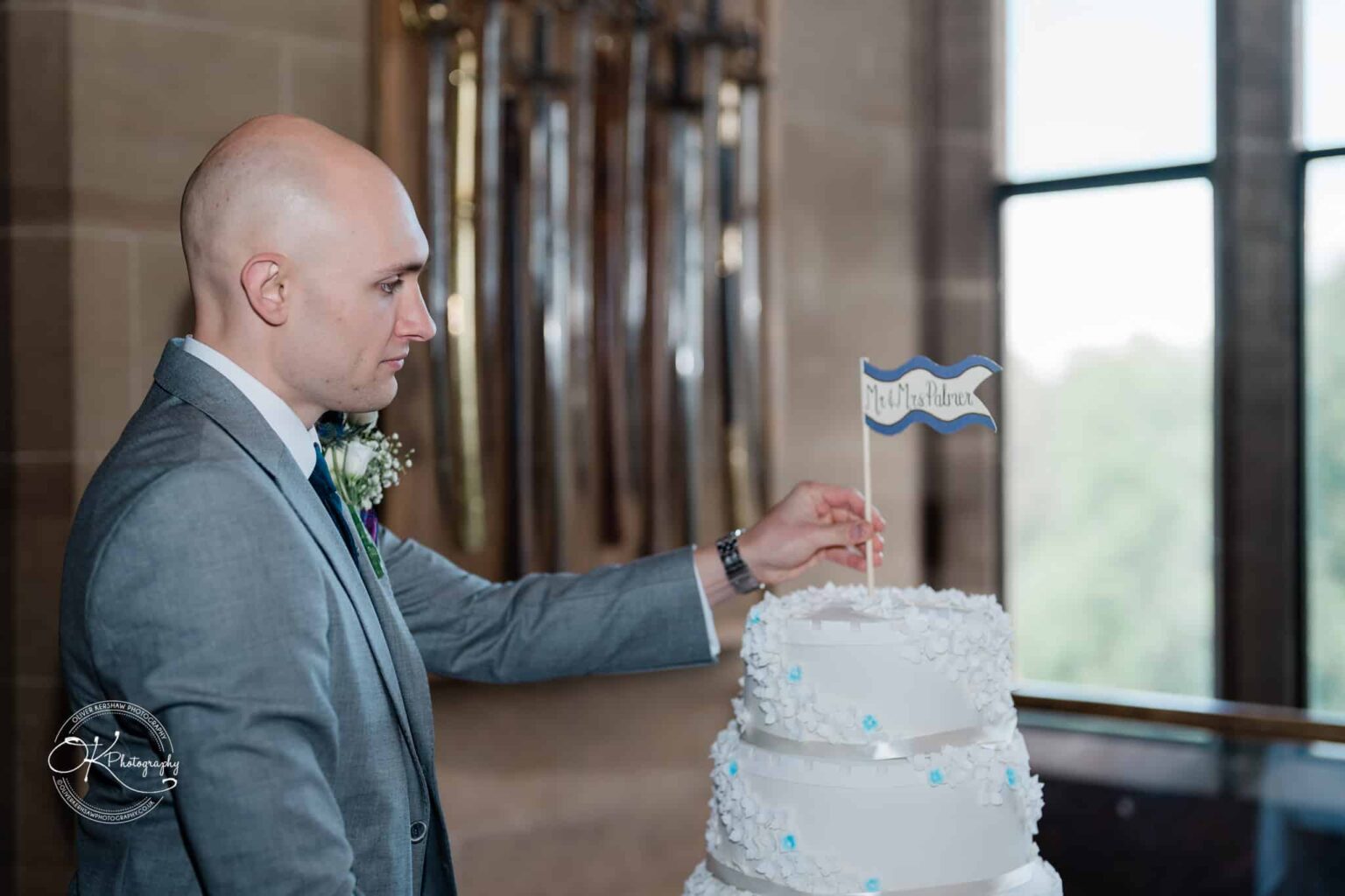 A man in a grey suit places a small flag with the text "Mr. & Mrs. Palmer" on a white tiered wedding cake at Warwick Castle.