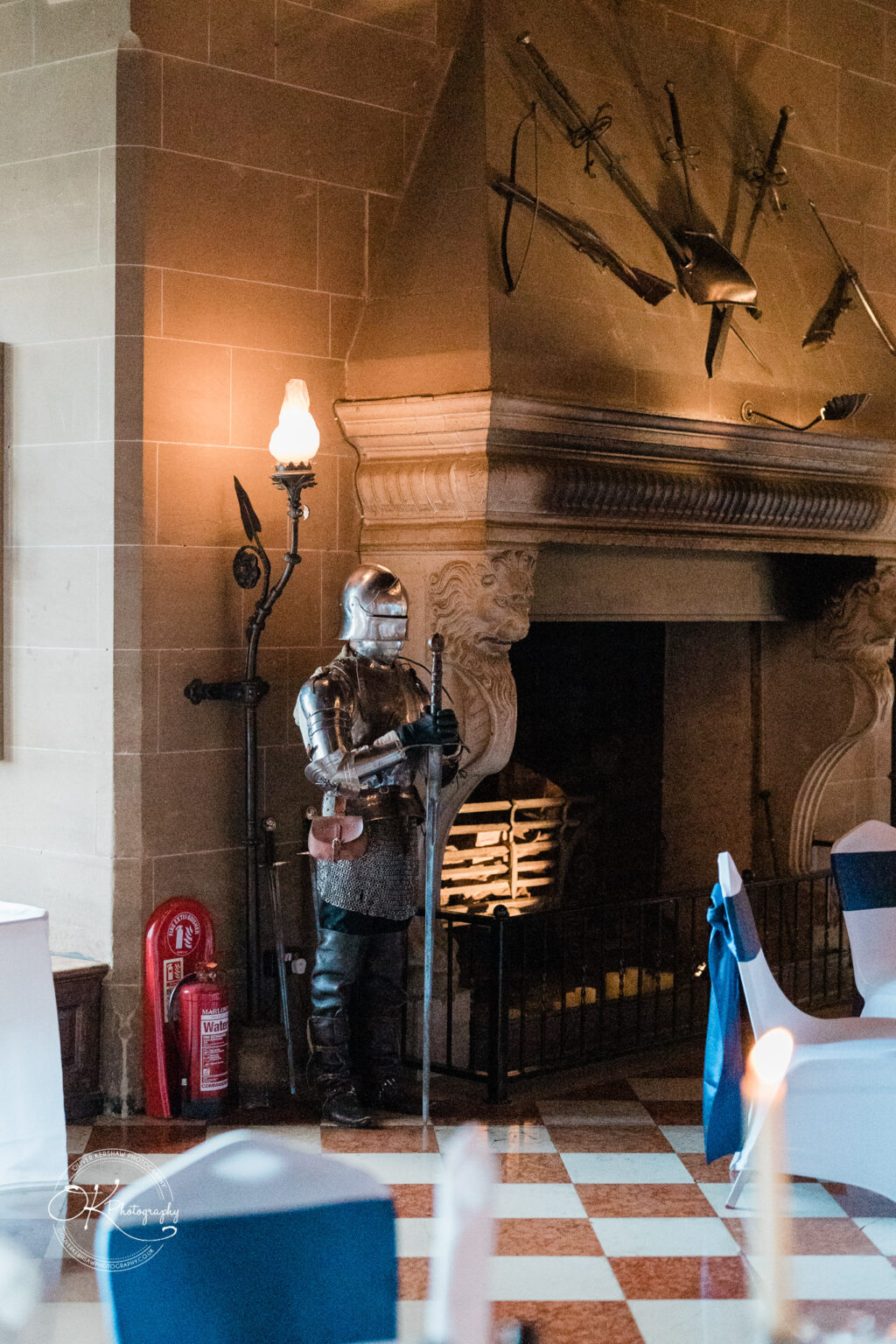 Armoured knight statue beside a large stone fireplace, with medieval weapons mounted above, located in a room at Warwick Castle.