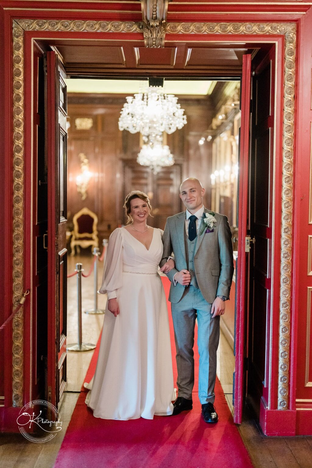 A bride and groom standing arm-in-arm at an ornate doorway in Warwick Castle, adorned with chandeliers and elaborate decor.