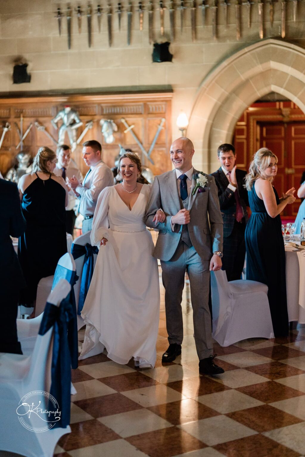 A newlywed couple smiling and walking arm-in-arm in an ornate hall with medieval armour and swords displayed on the walls, surrounded by applauding guests.
