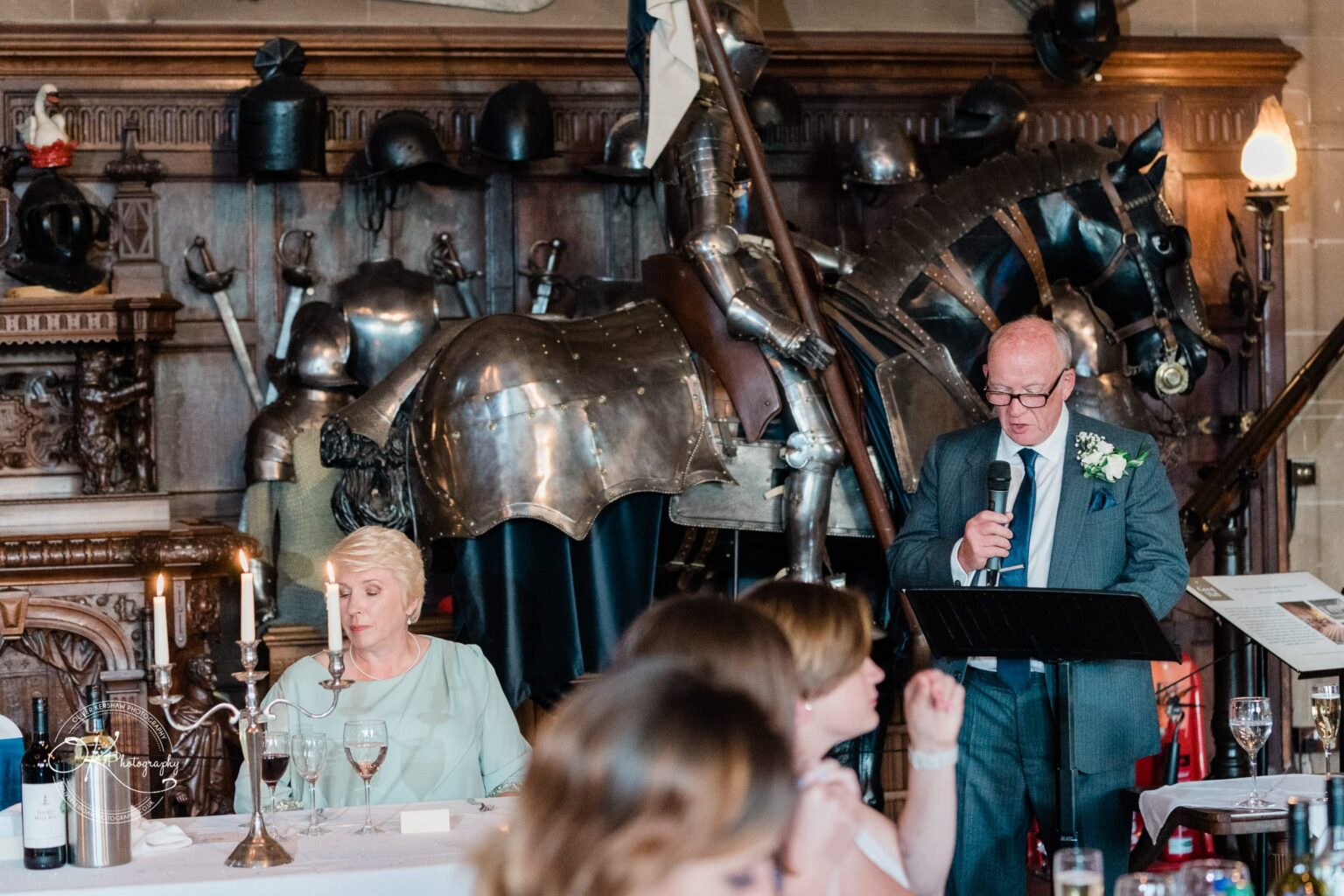 A man in a suit giving a speech with a microphone in front of a medieval knight's armour display at Warwick Castle; a woman is seated at a table with glasses of wine and a candlestick.