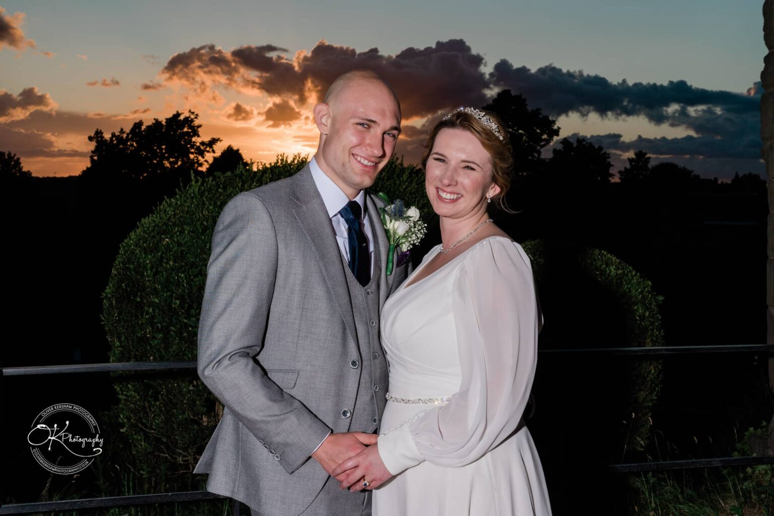 A bride and groom posing together in front of a sunset with greenery in the background.