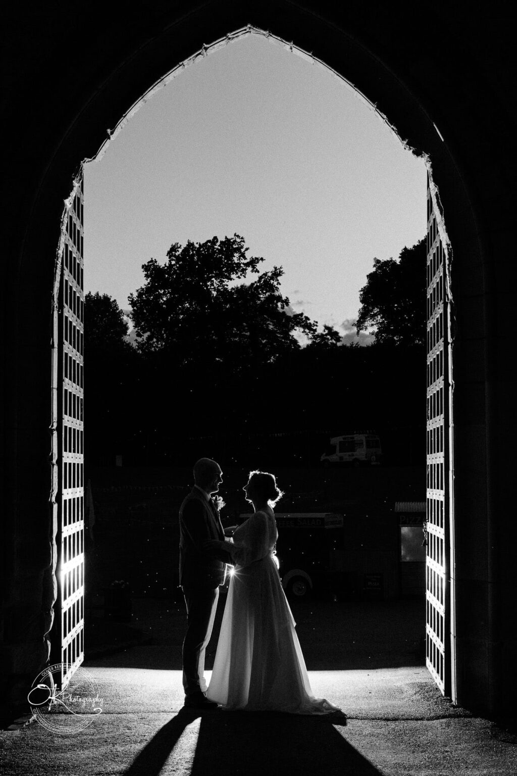 Silhouette of a couple in wedding attire standing in the arched entrance of Warwick Castle with trees visible in the background.