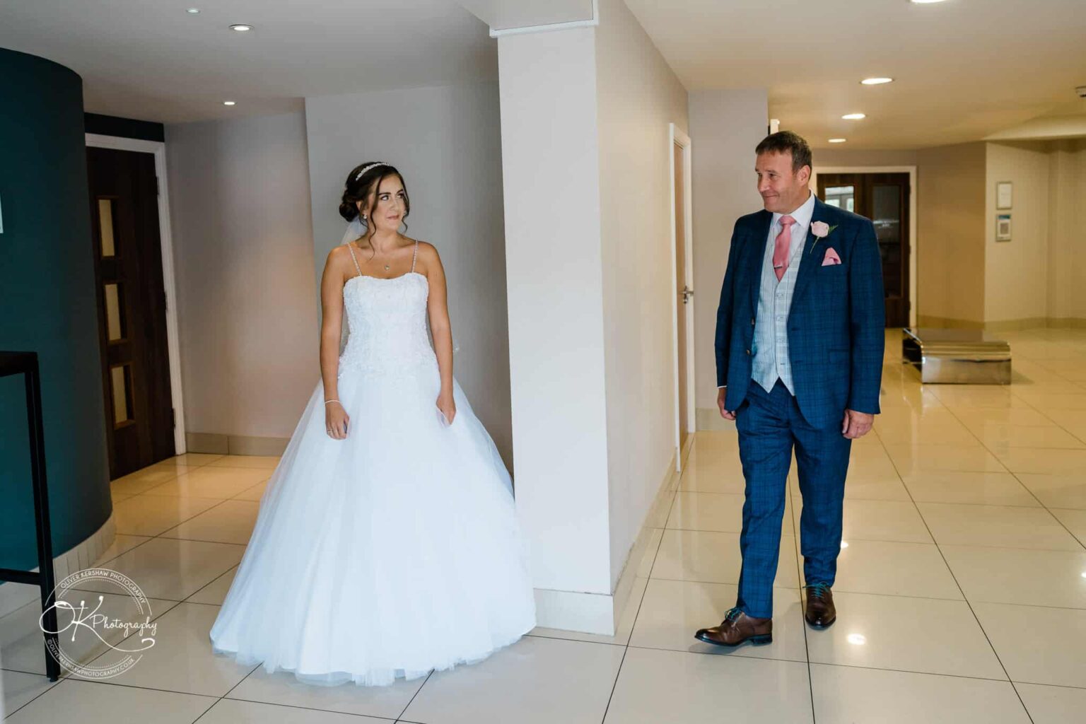 Warwick House Wedding Photography Bride in a white wedding dress looking at groom in a blue suit and pink tie in a hallway.