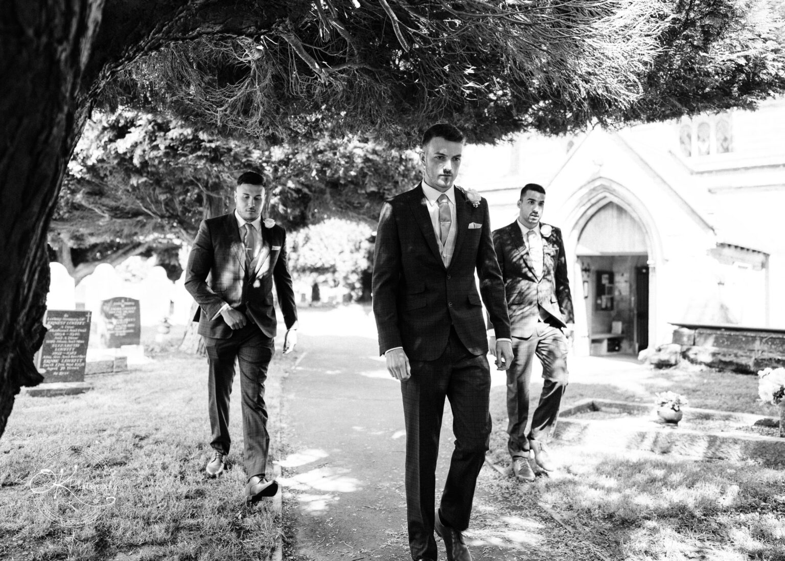 Warwick House Wedding Photography Three men in suits walking towards the camera under a large tree next to a church building.