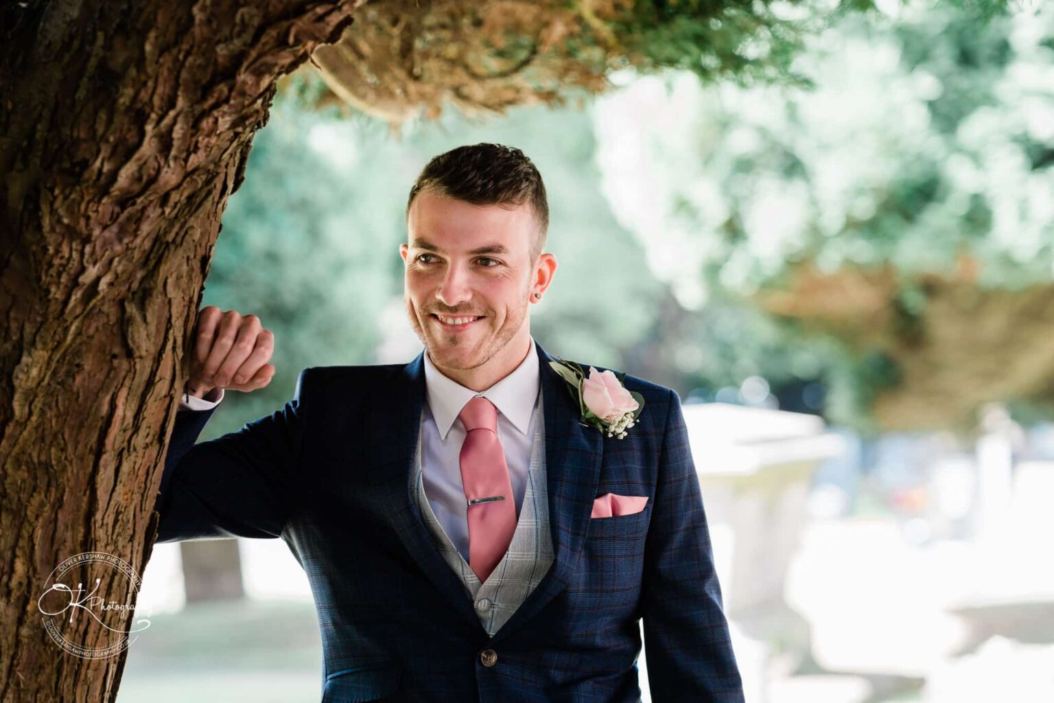Warwick House Wedding Photography Groom in a blue suit with pink tie and boutonniere leaning against a tree, smiling.