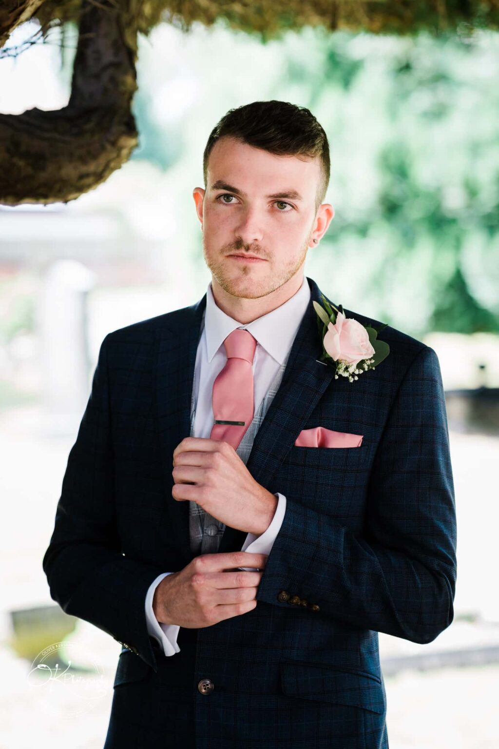 Warwick House Wedding Photography A man in a formal suit with a pink tie and a boutonnière adjusts his cufflink, standing outdoors with greenery in the background.