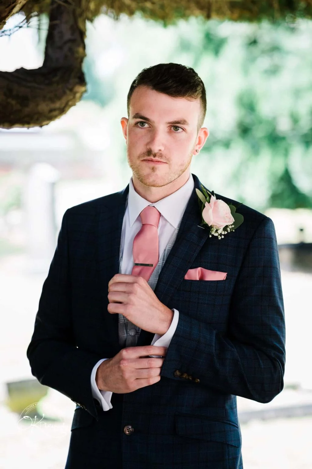 Warwick House Wedding Photography A man in a formal suit with a pink tie and a boutonnière adjusts his cufflink, standing outdoors with greenery in the background.