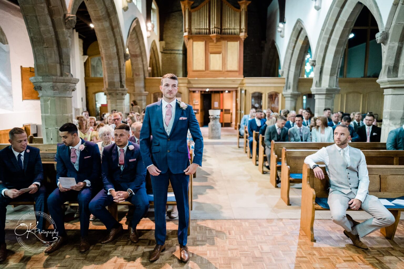 Warwick House Wedding Photography A wedding ceremony inside a church with groomsmen wearing blue suits and pink ties, and a man sitting on the right in light-coloured attire. Guests are seated in the pews, and the church features stone arches and wooden elements.