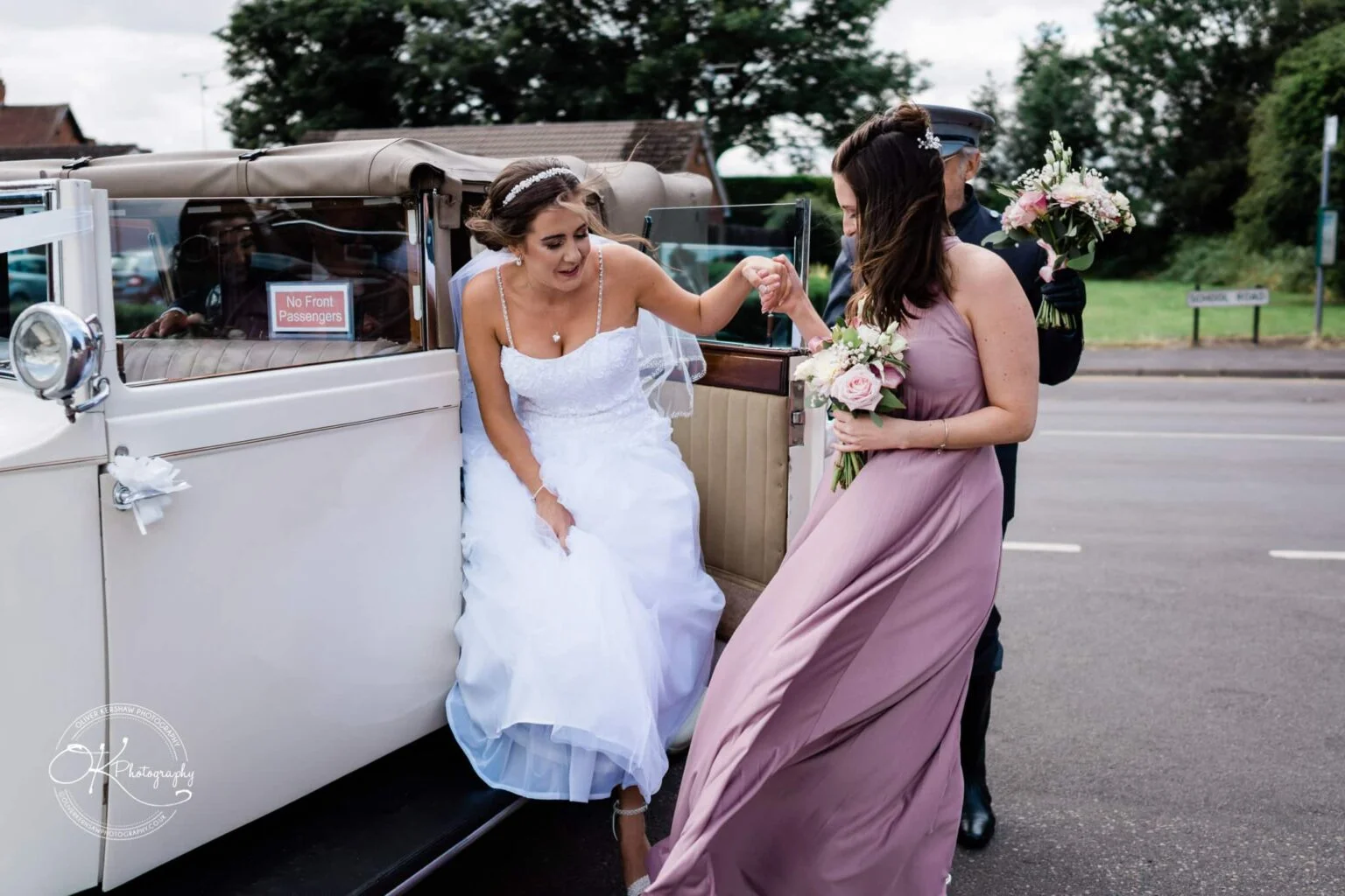 Warwick House Wedding Photography A bride in a white gown steps out of a vintage white car, assisted by a woman holding a bouquet.