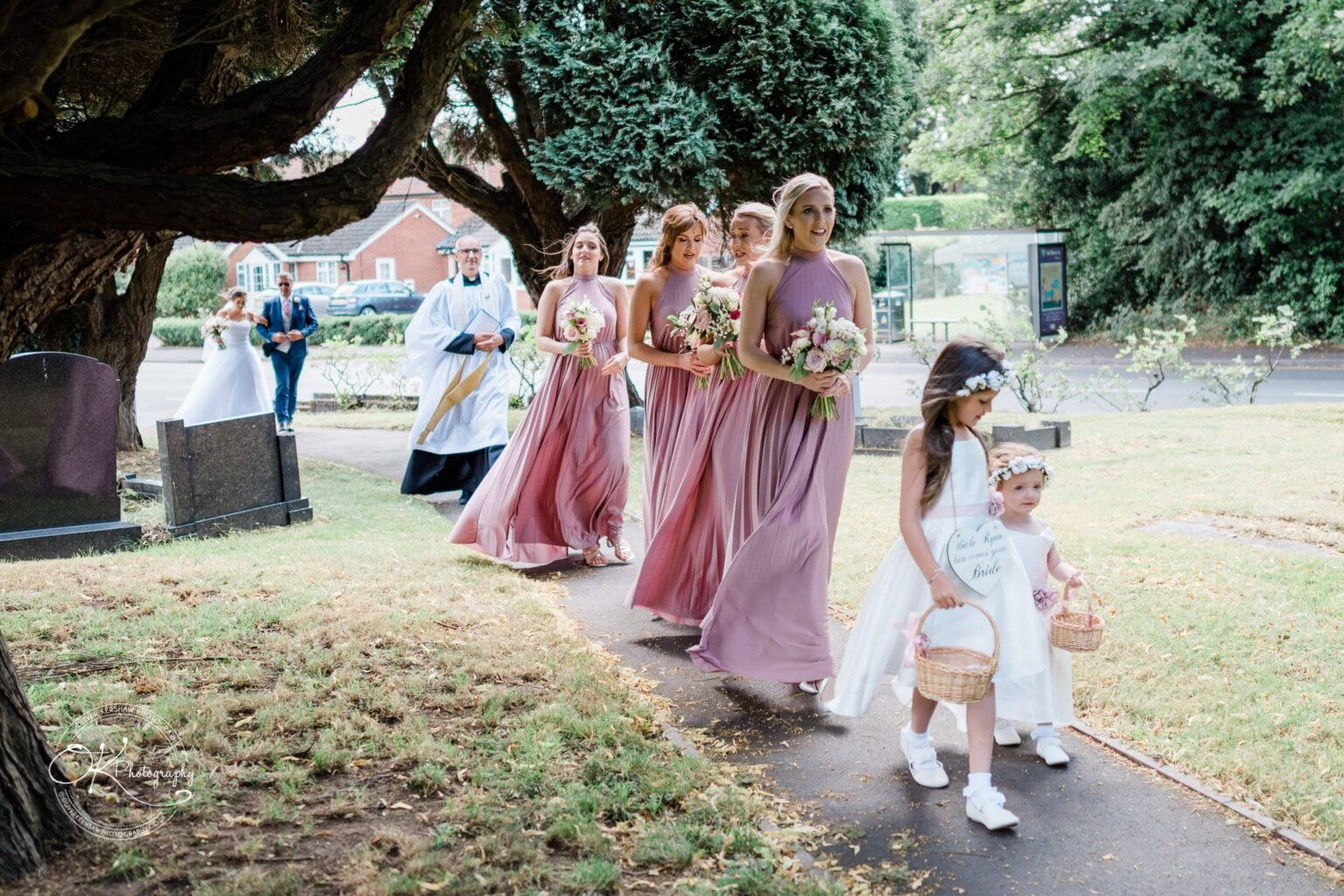 Warwick House Wedding Photography A wedding procession with bridesmaids in lavender dresses and flower girls in white, walking along a pathway near a church, led by a clergyman.
