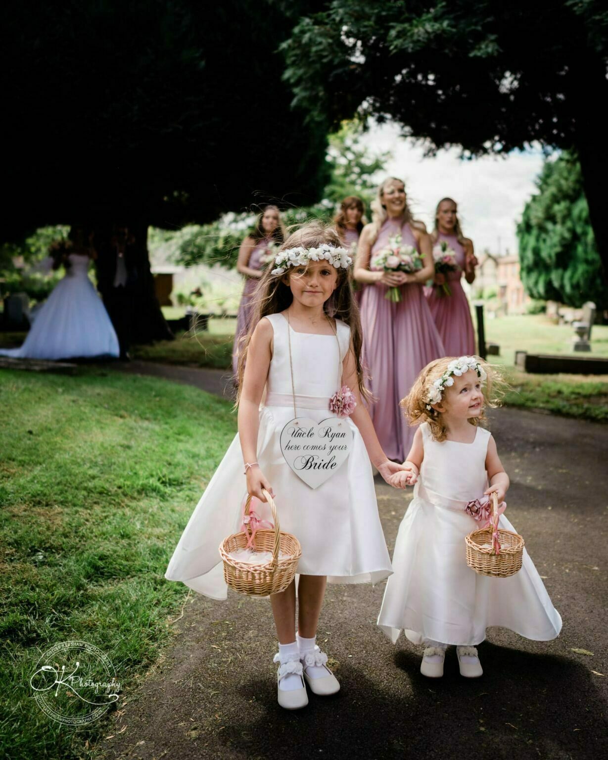 Warwick House Wedding Photography Two flower girls in white dresses and floral headbands walk hand-in-hand, holding baskets, with bridesmaids and the bride in the background.