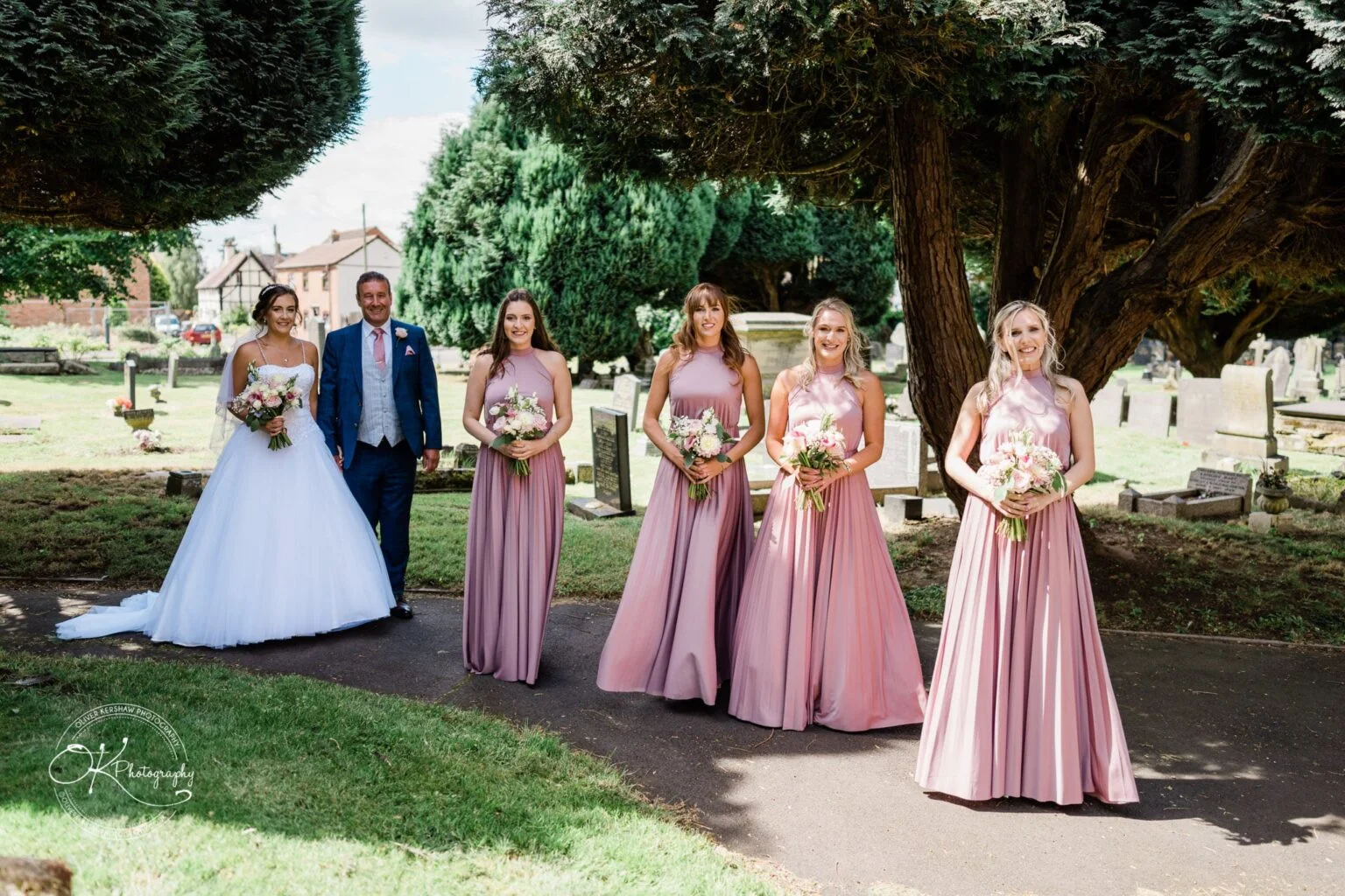 Warwick House Wedding Photography Bride in white gown with groom and four bridesmaids in pink dresses holding bouquets, standing outdoors by trees and gravestones at Warwick House.