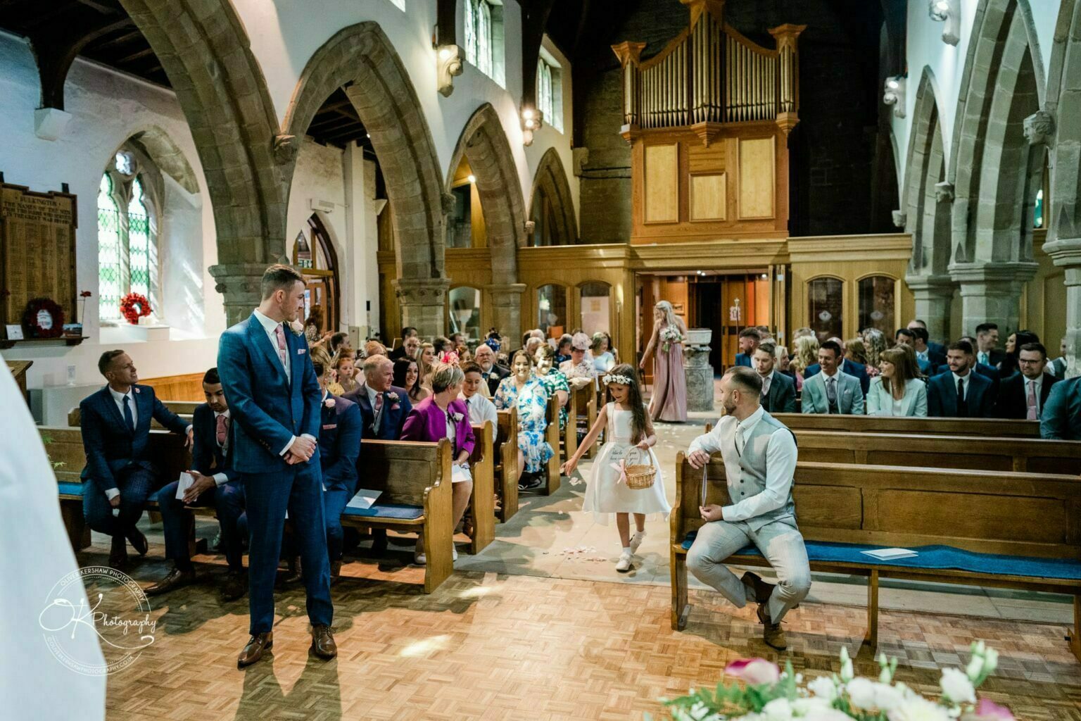 Warwick House Wedding Photography Wedding ceremony in a church with guests seated in wooden pews and a flower girl walking down the aisle.