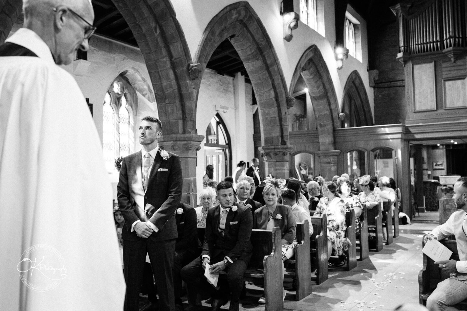 Warwick House Wedding Photography Black and white photo of a wedding ceremony inside a church, with the groom standing in the aisle and guests seated in pews on either side.