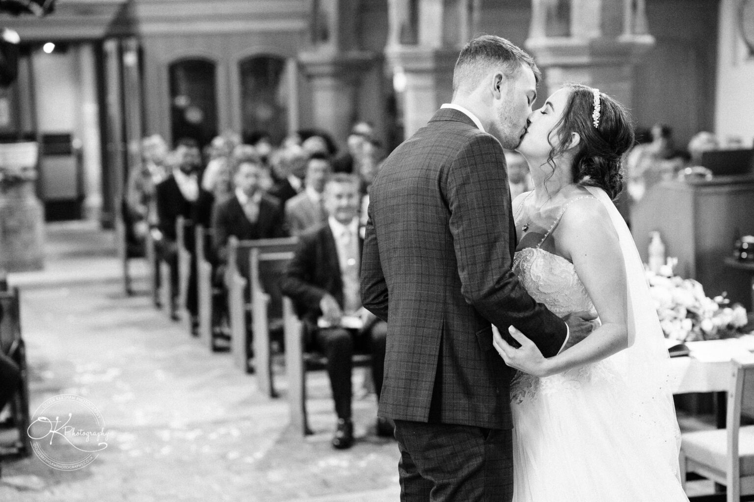 Warwick House Wedding Photography Bride and groom in wedding attire share a kiss in front of seated guests inside a church.