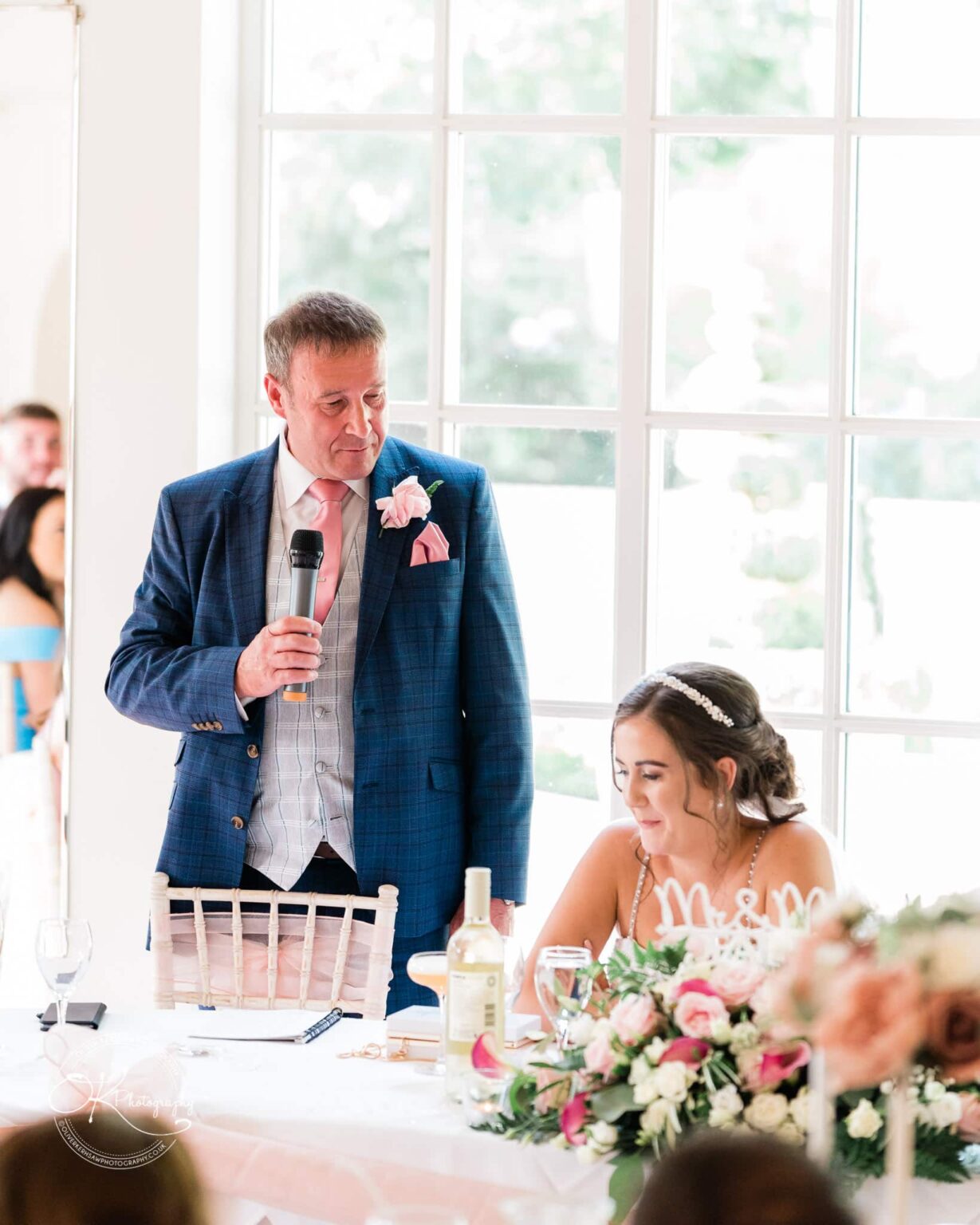 Warwick House Wedding Photography A man in a blue suit and pink tie is giving a speech at a wedding reception, while the bride sits next to him smiling.