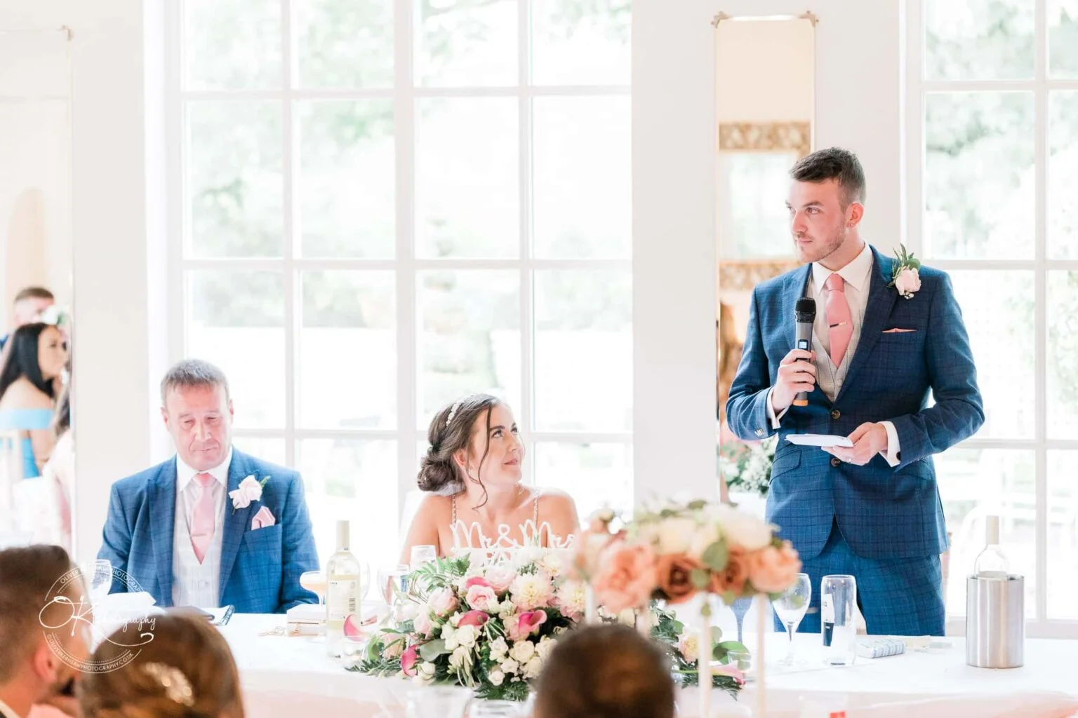 Warwick House Wedding Photography A wedding reception scene at Warwick House with a man giving a speech, the bride and another man seated at the table adorned with flowers.