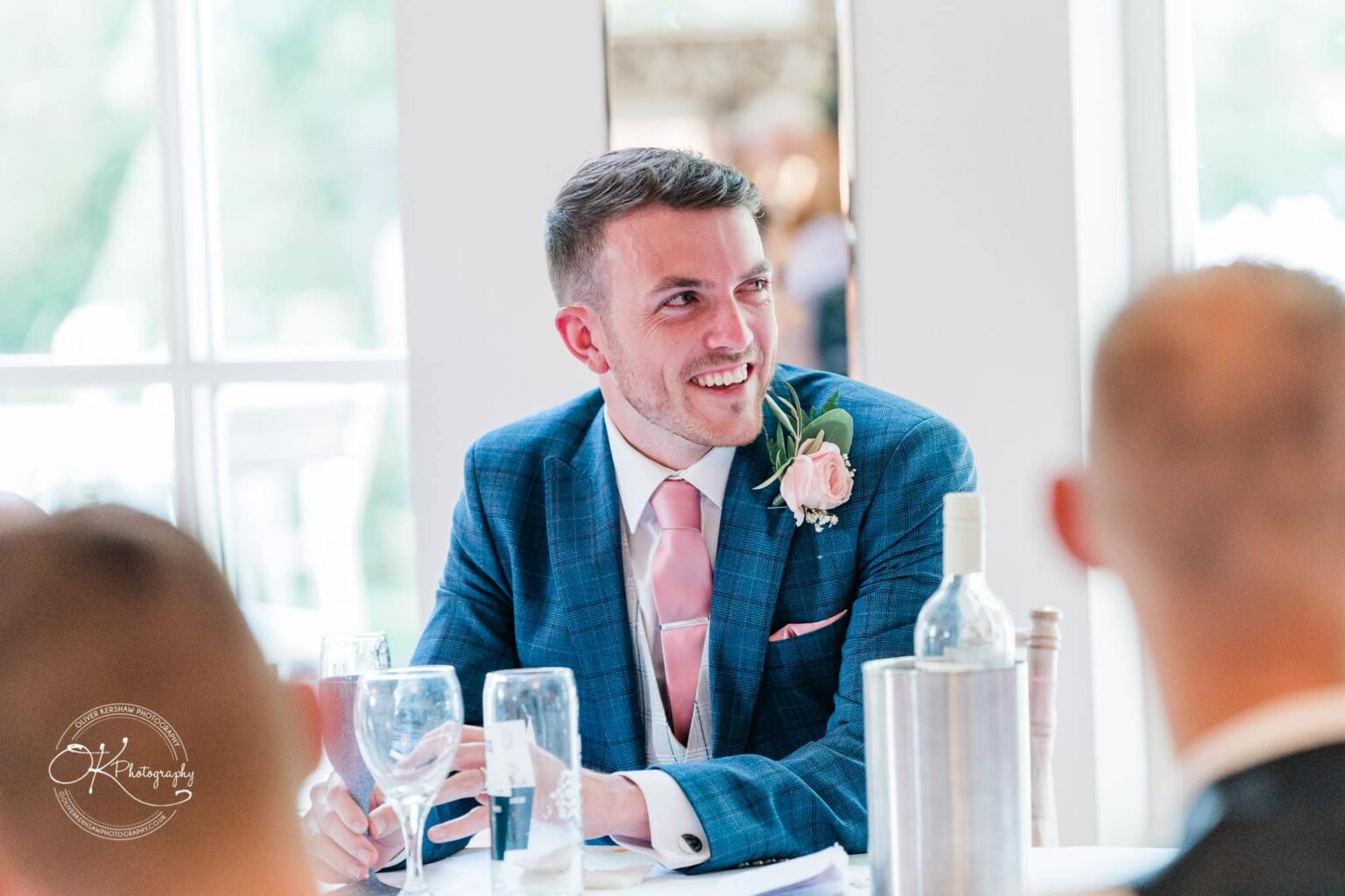 Warwick House Wedding Photography A man in a blue suit with a pink tie smiling and sitting at a table during a wedding reception.