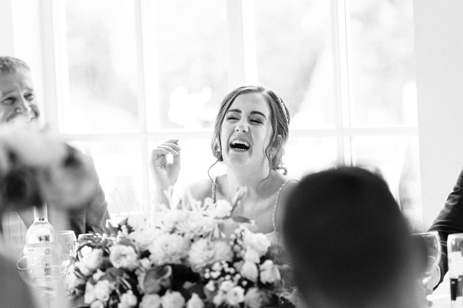 Warwick House Wedding Photography A woman in a wedding dress laughs joyfully at a table adorned with flowers, with other people and a window in the background.